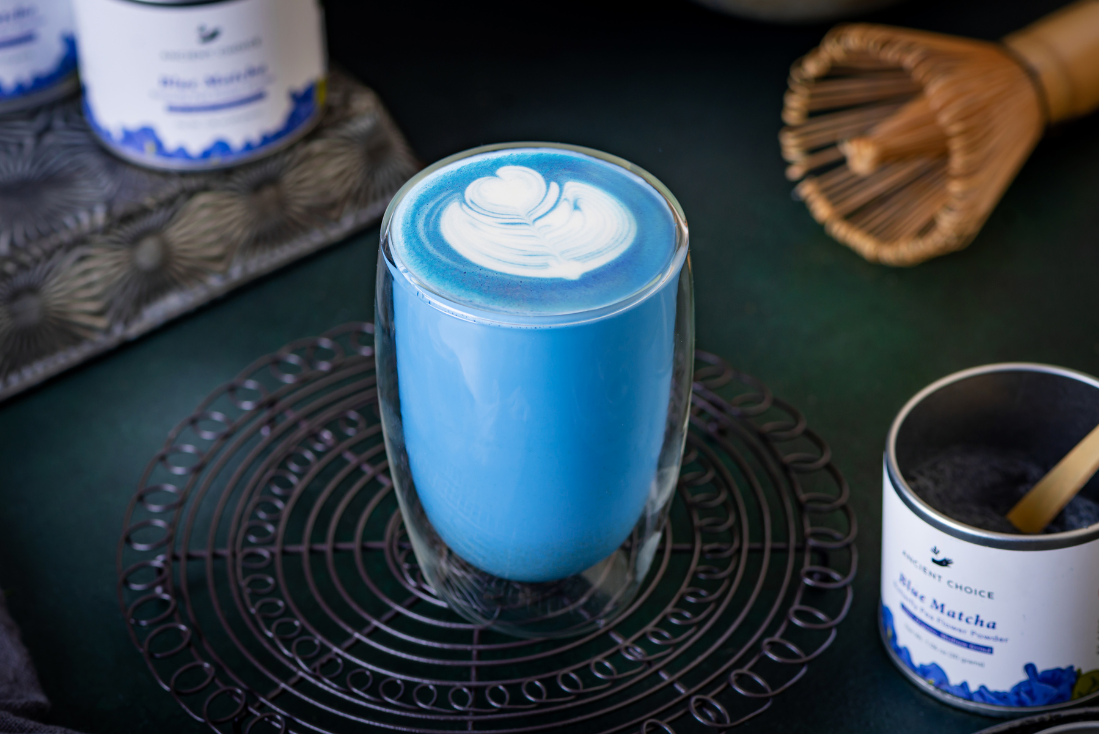 A cup of blue matcha latte with latte art surrounded by Ancient Choice Blue Matcha tin canisters