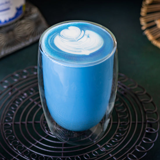 A glass of blue matcha latte with latte art surrounded by Ancient Choice Blue Matcha tin canisters