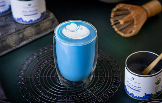 A cup of blue matcha latte with latte art surrounded by Ancient Choice Blue Matcha tin canisters