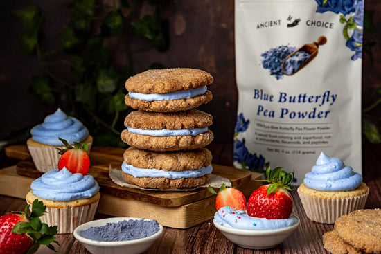 3 sandwich cookies with blue frosting, stacked on top of each other. Next to them, a cupcake with blue frosting and a strawberry as decoration. Behind, a butterfly pea flower powder bag.