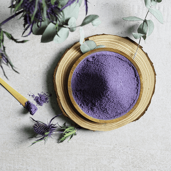 Extra fine blue matcha powder with spoon, in a wooden bowl on rustic wood slice, surrounded by leaves.