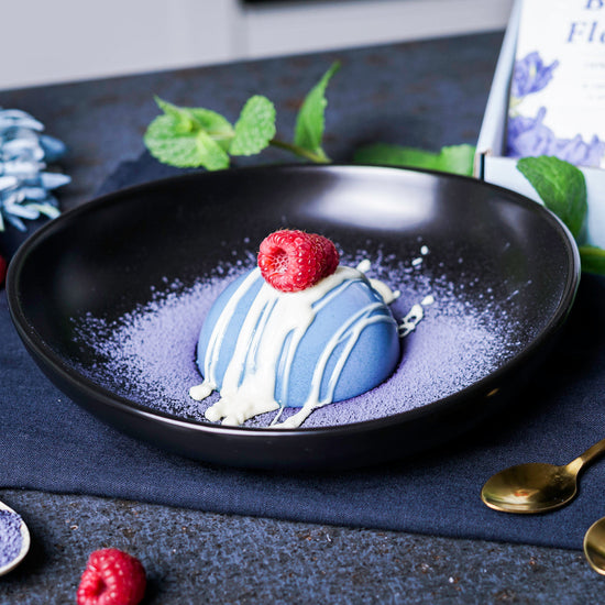 Butterfly pea flower mousse, garnished with butterfly pea flower powder, raspberries, and white cream, served on a black plate. In the background, a bag of butterfly pea flower powder.