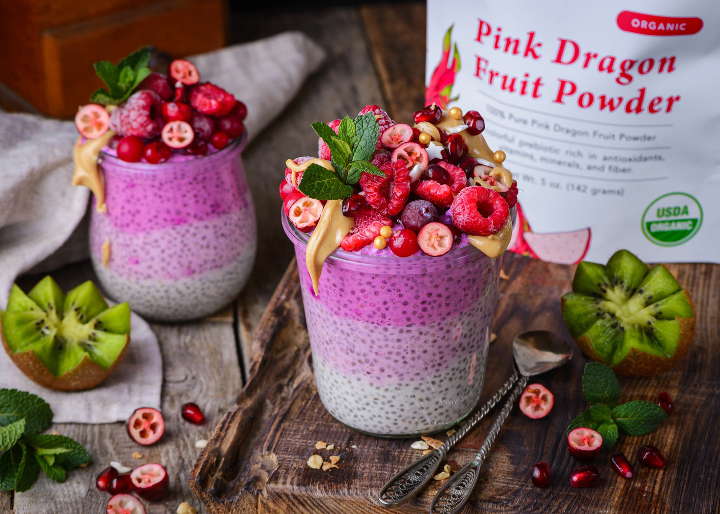 Two jars of layered chia seed pudding topped with fresh raspberries, pomegranate seeds, and mint, alongside a bag of Ancient Choice Pink Dragon Fruit Powder on a wooden surface