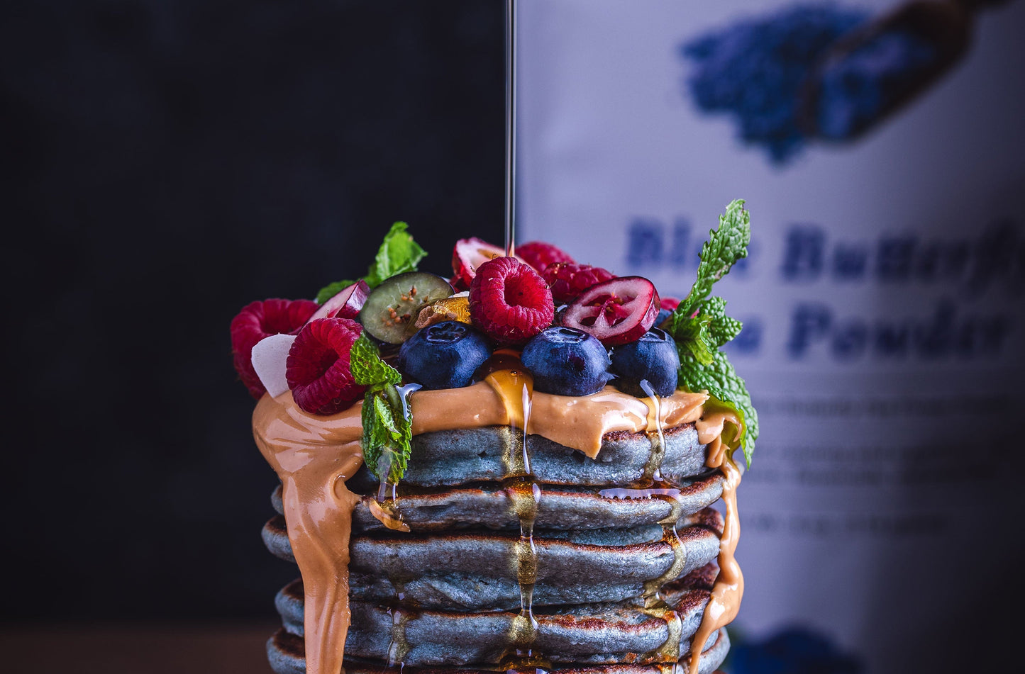 Tall stack of blueberry pancakes topped with peanut butter, berries, and mint, with syrup drizzling down