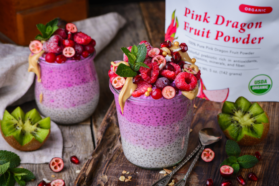 Two jars of layered chia seed pudding topped with fresh raspberries, pomegranate seeds, and mint, alongside a bag of Ancient Choice Pink Dragon Fruit Powder on a wooden surface