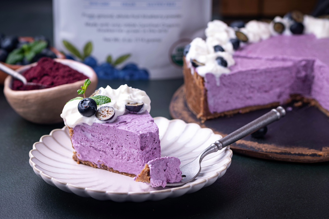 Slice of wild blueberry cheesecake topped with whipped cream, fresh blueberries, and mint, served on a plate with the full cake in the background.